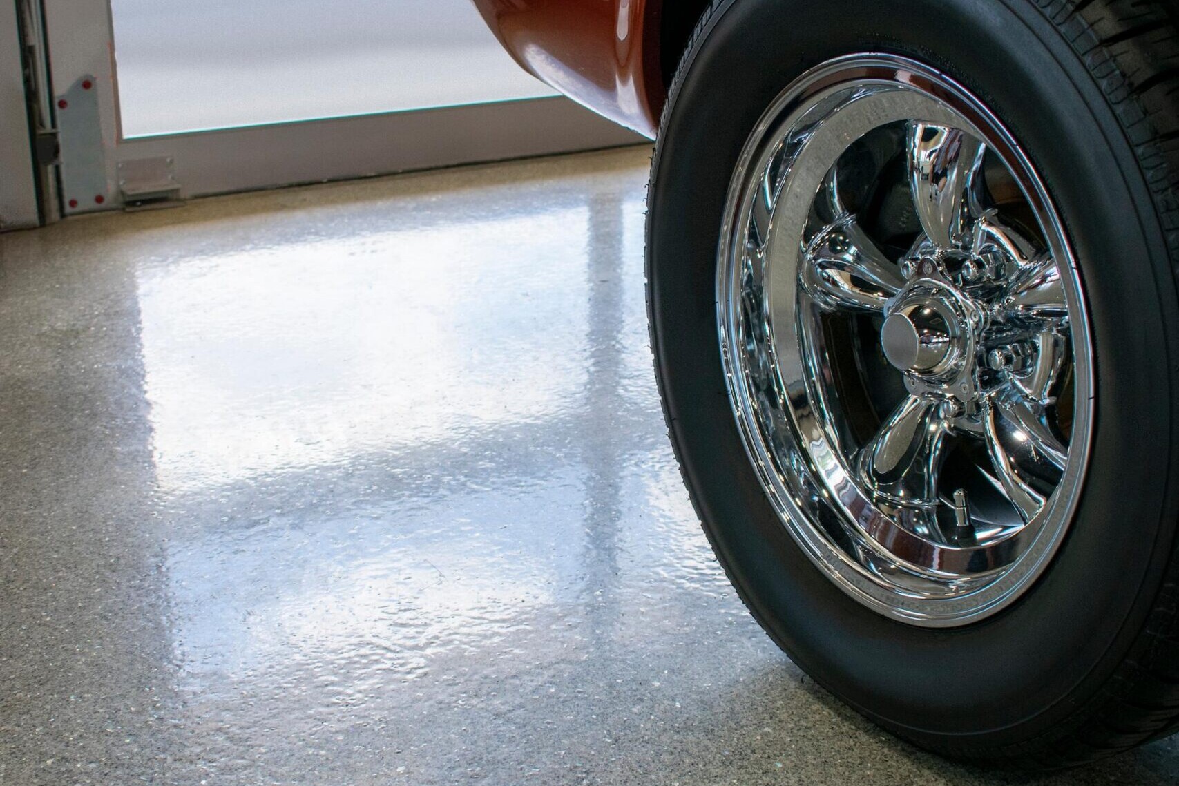 The rear wheel of an orange classic car parked on a shiny, speckled gray floor next to a glass garage door.