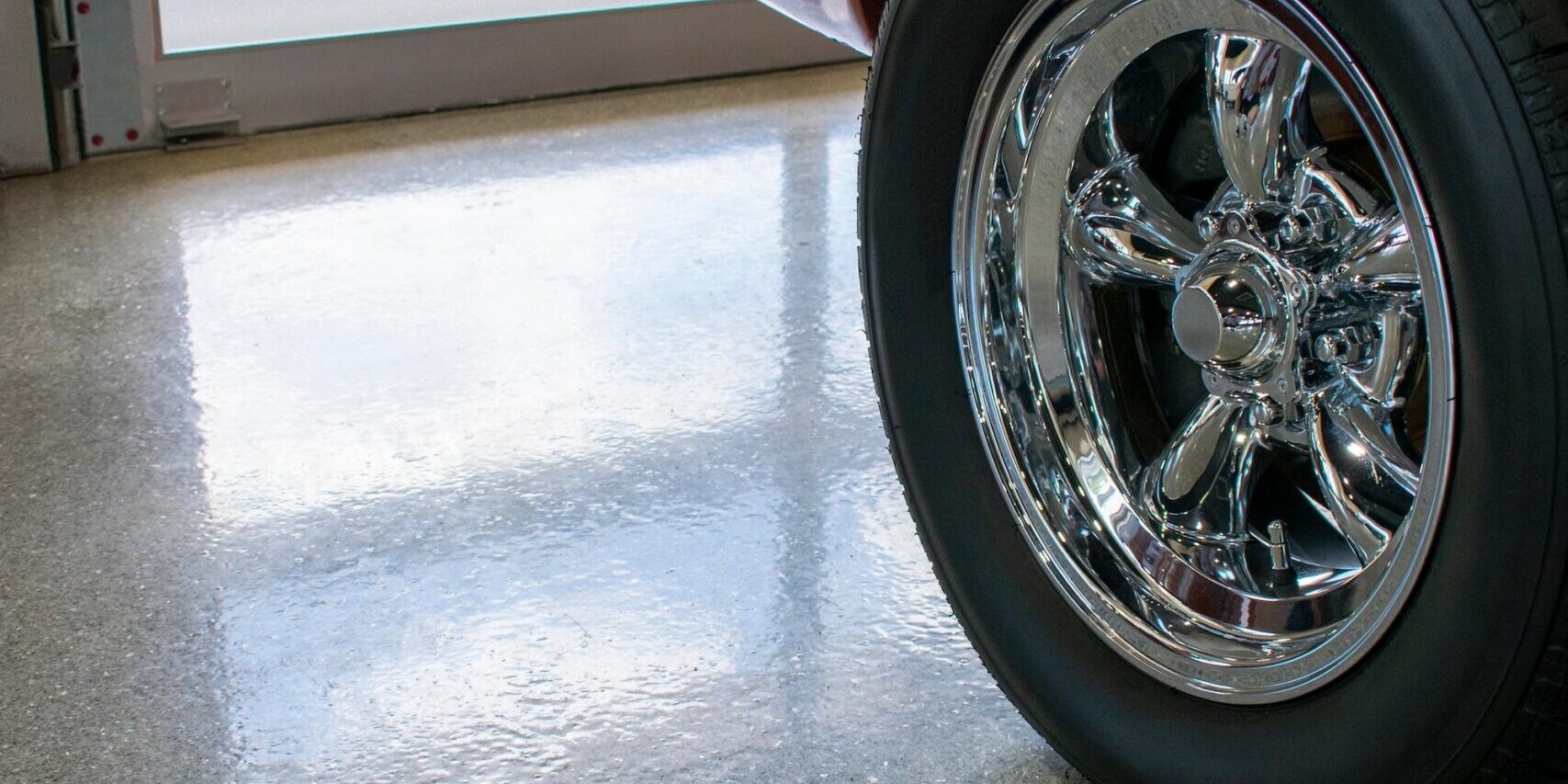 The rear wheel of an orange classic car parked on a shiny, speckled gray floor next to a glass garage door.