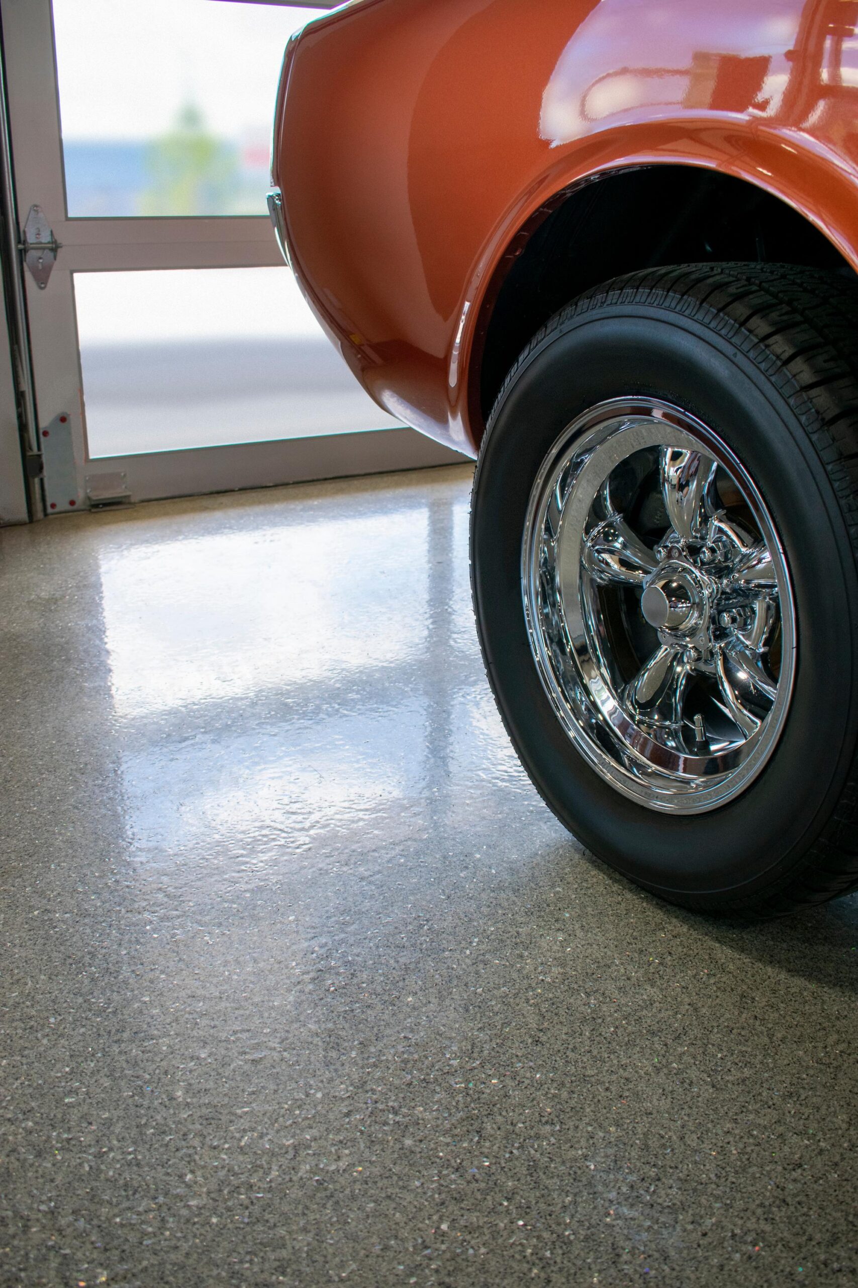 The rear wheel of an orange classic car parked on a shiny, speckled gray floor next to a glass garage door.