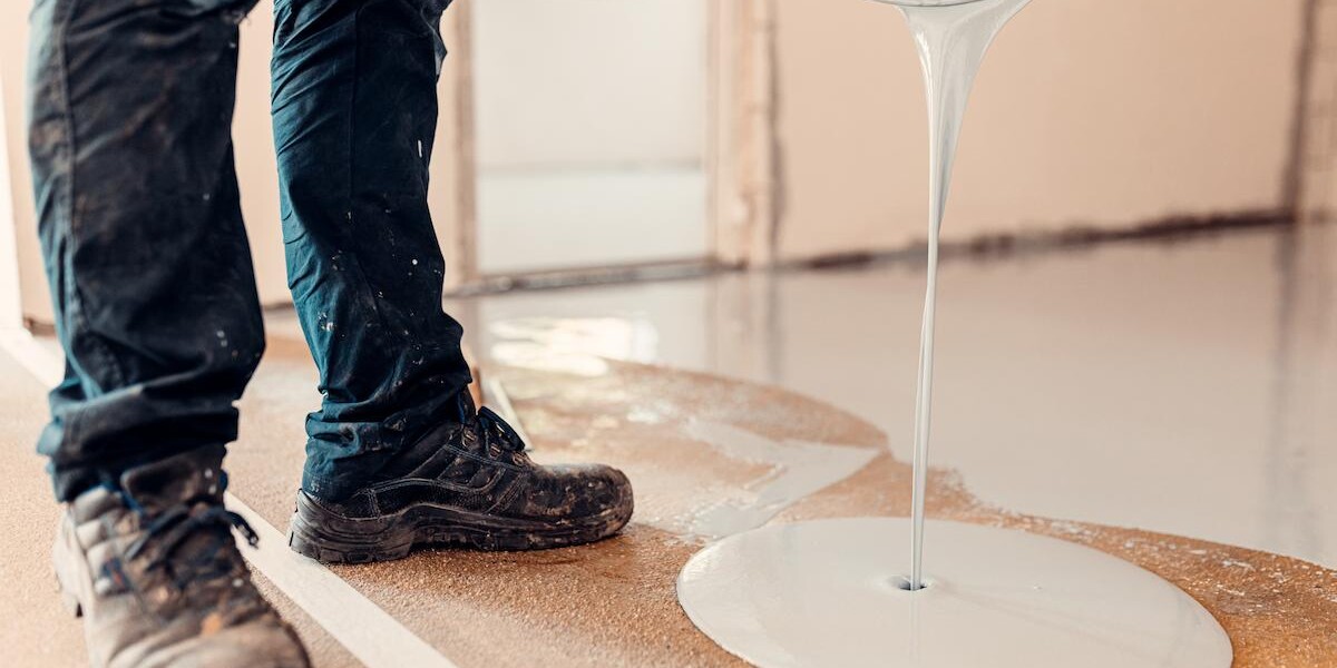 A person in work boots pours a thick, light gray liquid from a bucket onto an unfinished floor.