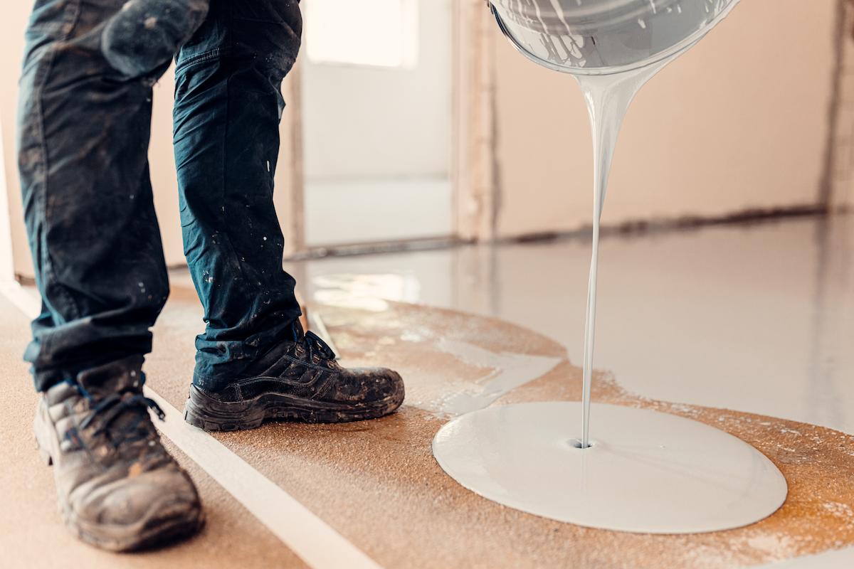 A person in work boots pours a thick, light gray liquid from a bucket onto an unfinished floor.