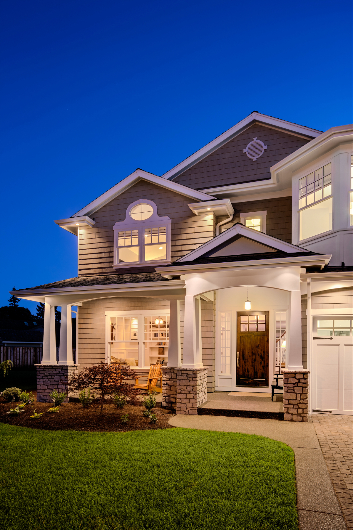 house with a covered front porch, stone pillars, landscaped lawn, and warm exterior lighting