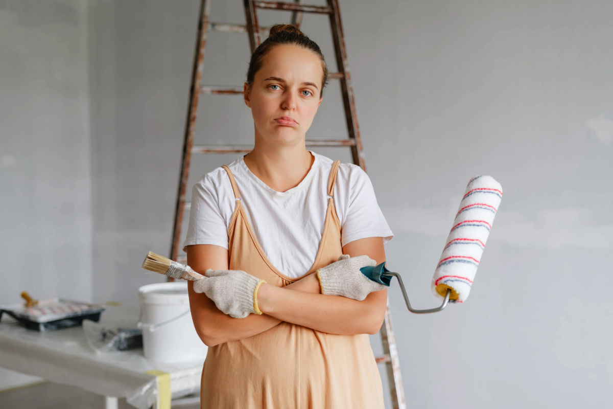 A woman is in an empty room holding a paint roller and a brush.